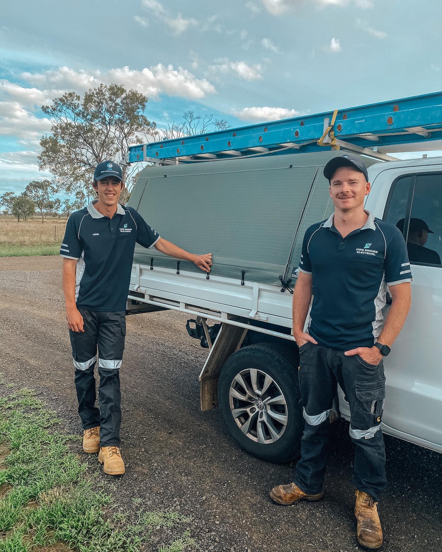 Two Chas Graham Electrical team members in branded uniforms standing beside a work ute with a ladder on the roof